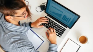 Overhead shot of a person reviewing their budget on a laptop with a notebook and coffee cup nearby, natural daylight, calm and focused expression, clean desk workspace